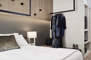 Quarto de hotel com cama, mesa de cabeceira, abajur, guarda-roupa e prateleira com sapatos.