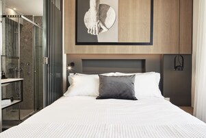 Quarto de hotel moderno com uma cama grande, chuveiro em cabine de vidro e decoração minimalista.