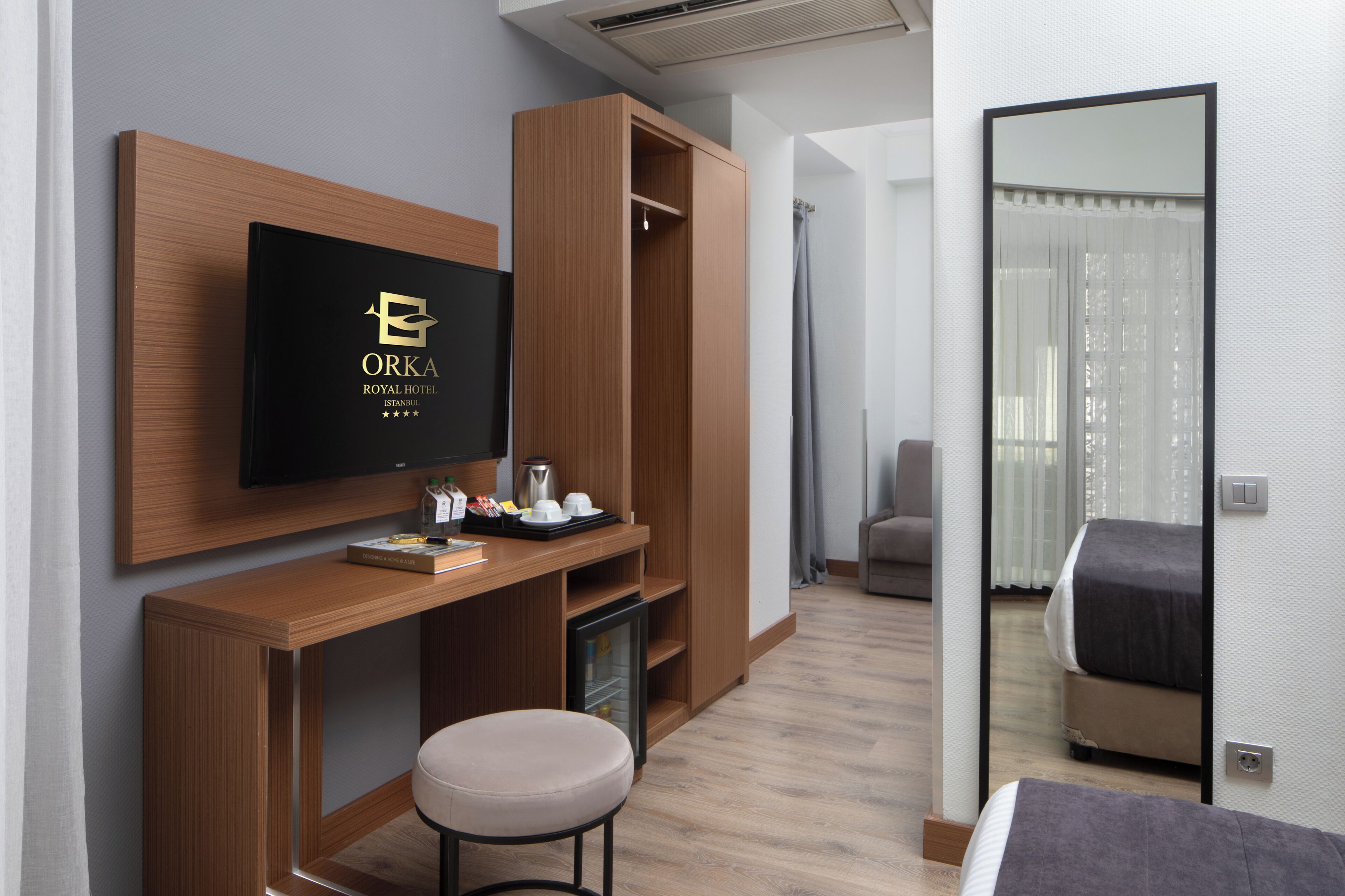 Moderni hotellihuone, jossa on puinen työpöytä, televisio, jossa näkyy hotellin nimi, minibaari ja sänky, josta avautuu näkymä kaupunkiin.