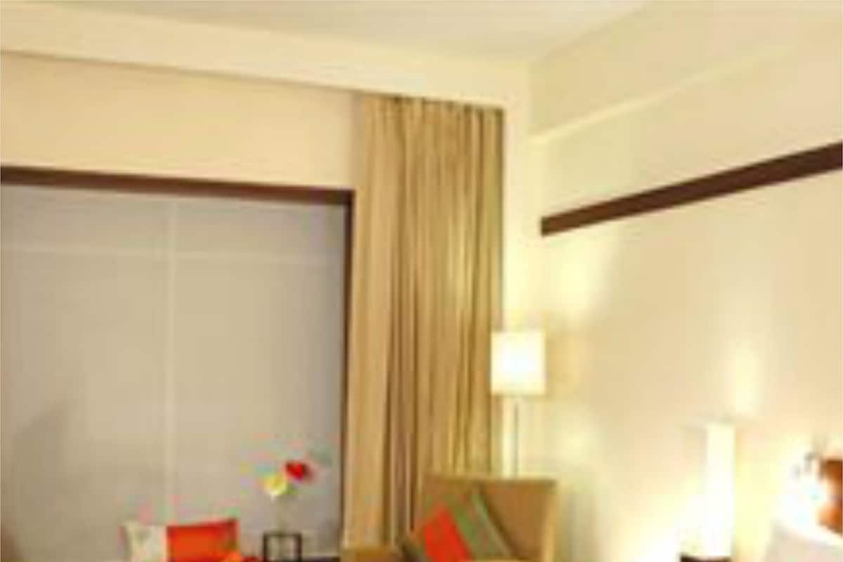 A hotel room with a bed, a chair, a small table with a lamp, and a window with curtains.