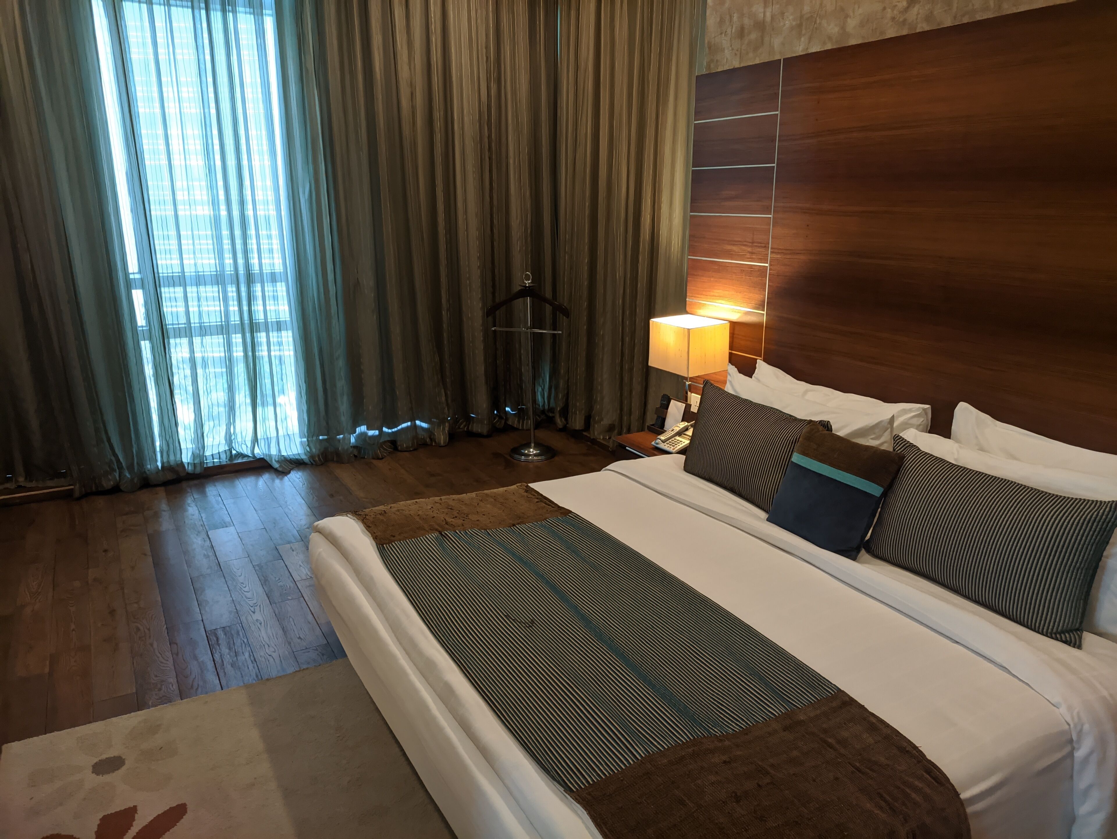 A hotel room with a large bed, a bedside table with a lamp, a wooden headboard, and a view of the outdoors through sheer curtains.