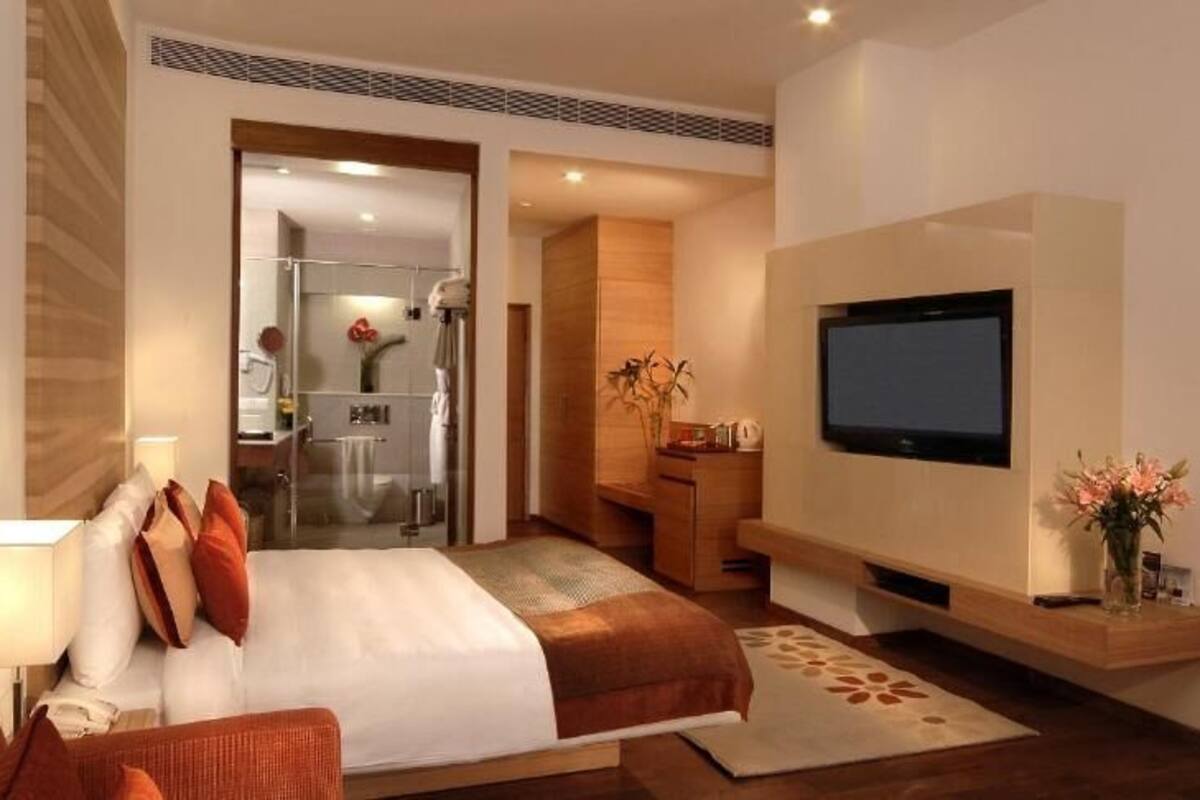A modern hotel room with a large bed, a flat-screen TV, a desk with a lamp, and a bathroom visible through an open door.