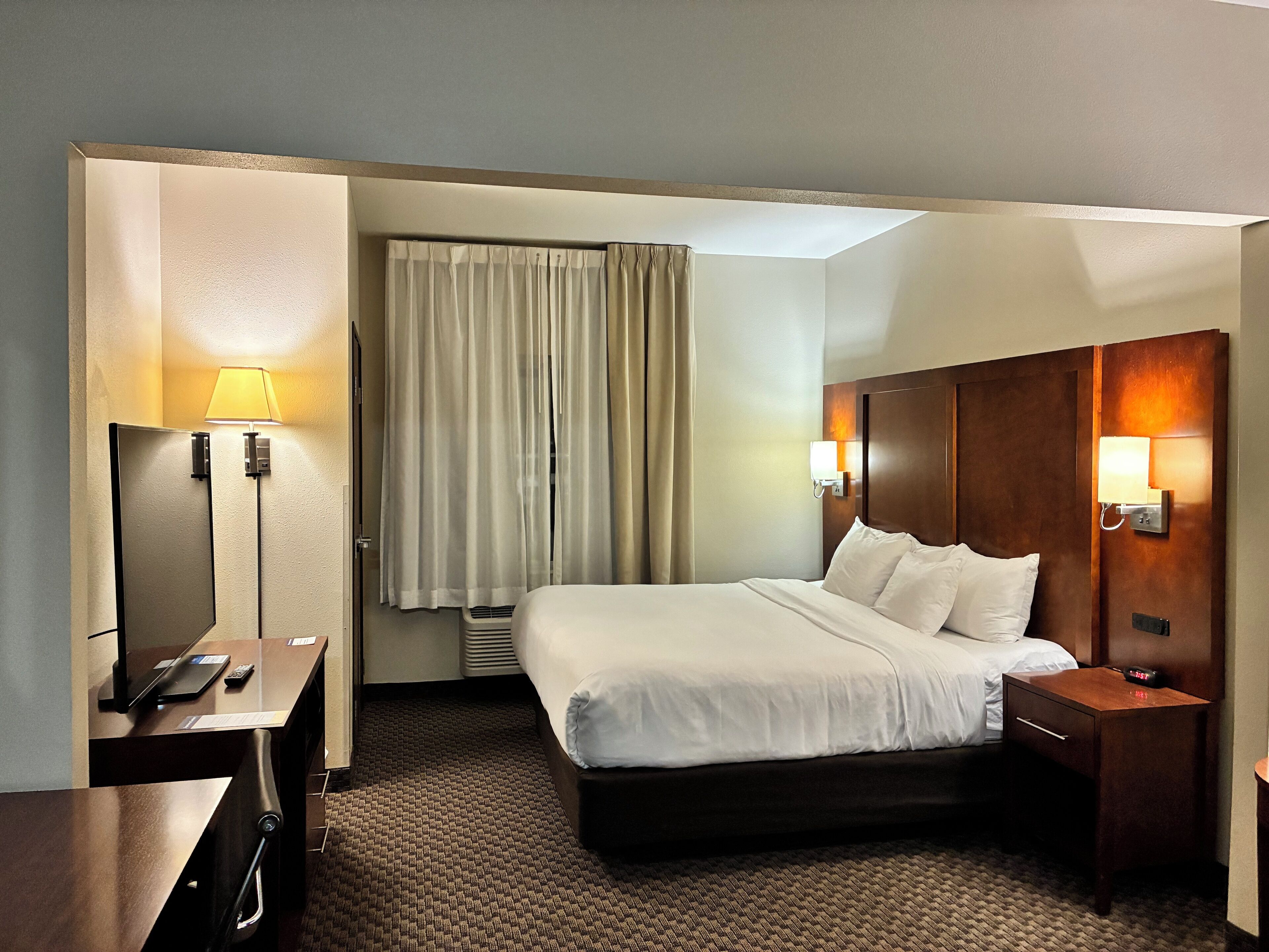 Une chambre d’hôtel avec un lit, des tables de chevet, un bureau, une lampe et une fenêtre avec des rideaux.