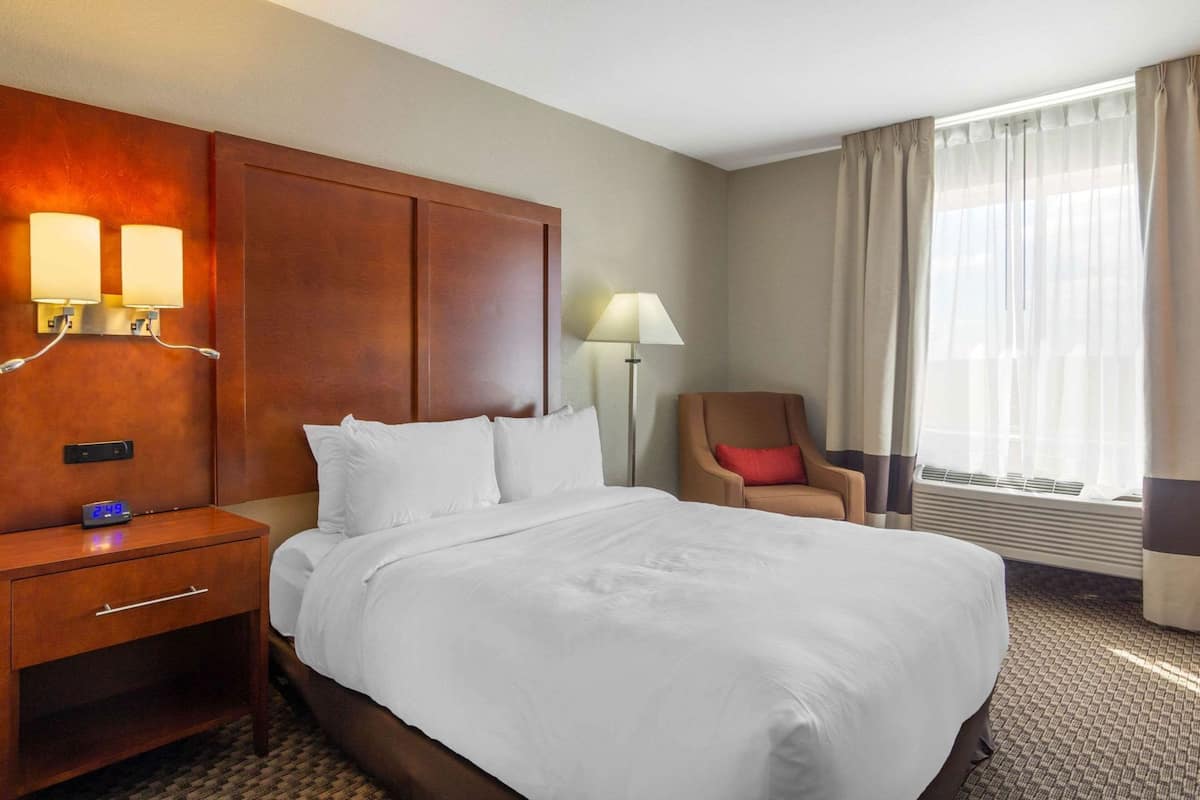 A hotel room with a bed, a nightstand with a clock, a chair, a lamp, and a window with curtains.