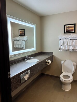 A bathroom with a granite countertop, a white sink, a mirror with a light, a toilet, and a towel rack with towels.