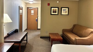 A hotel room with a brown leather sofa, a wooden bench, a small wooden table, and a white bed. There is a door leading to another room and a wall with two framed pictures. - Comfort Inn & Suites Sterling (Sterling)
