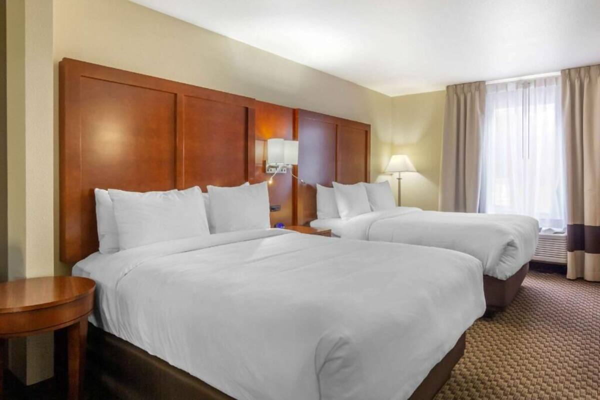 A hotel room with two beds, a wooden headboard, a bedside table, a lamp, and a window with curtains.