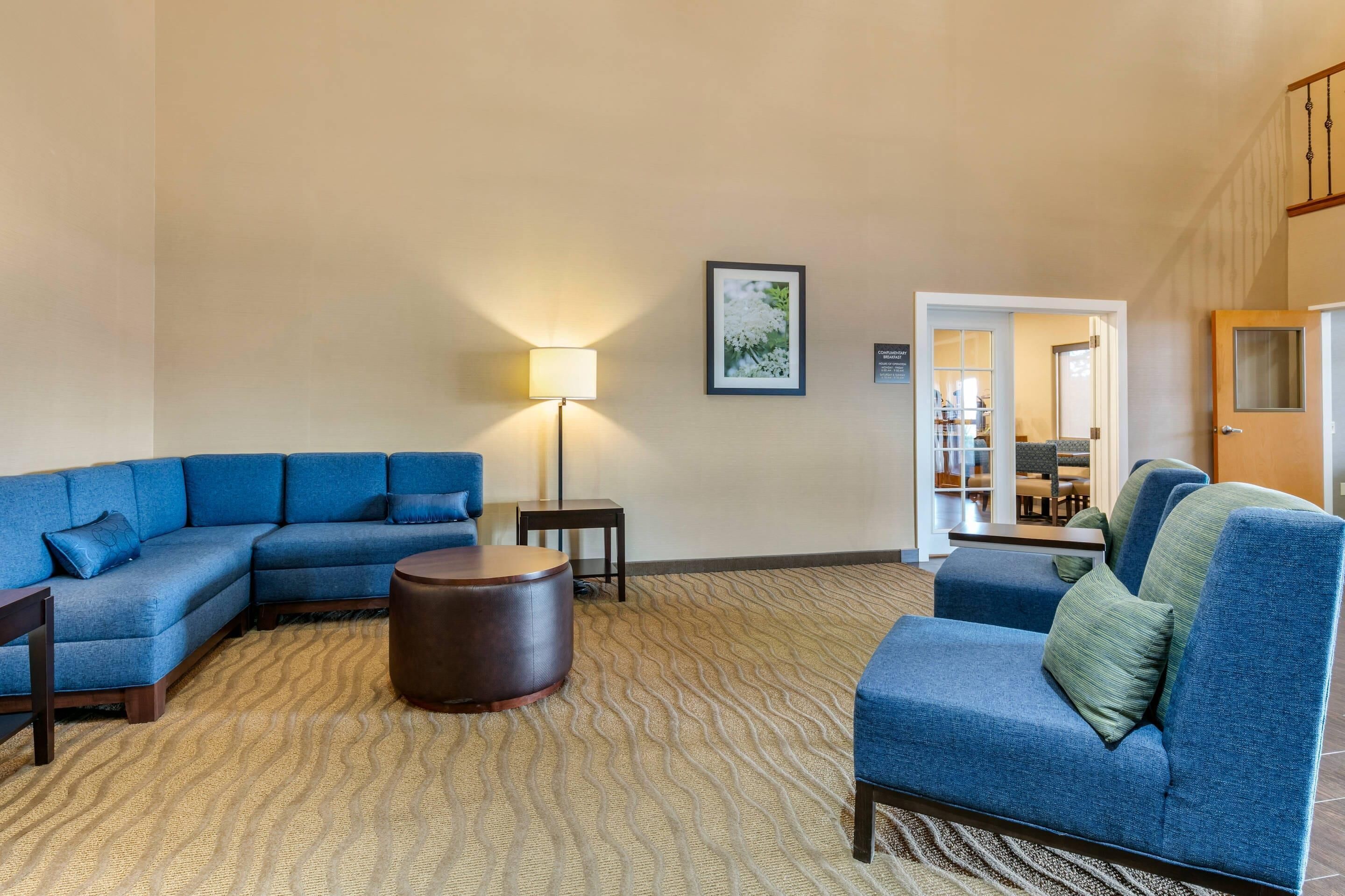 A living room with a sectional sofa, a round coffee table, and a lamp. There is a framed picture on the wall and a door leading to another room.