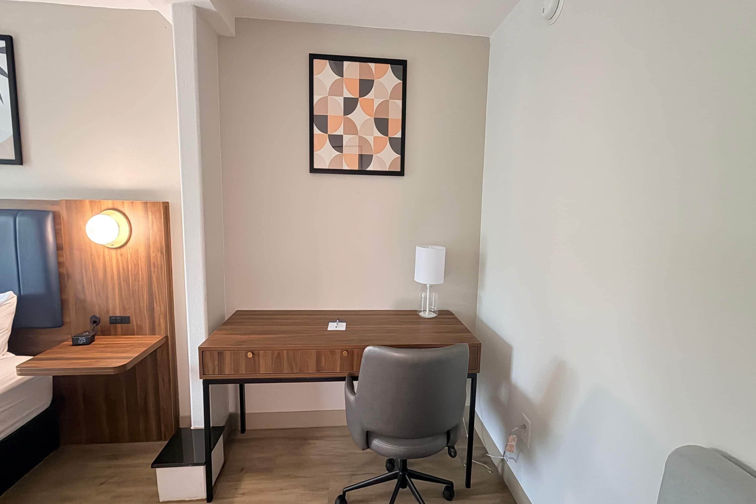 Et hotelværelse med et træskrivebord, en grå kontorstol, et natbord med en lampe og et billedværk på væggen.