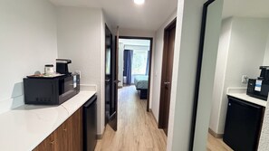 A narrow hallway with a kitchenette on one side, a bed in the adjacent room, and a view of the outdoors through a window.