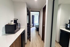 A narrow hallway leading to a bedroom with a bed and a window. There are two black coffee makers on the counter.