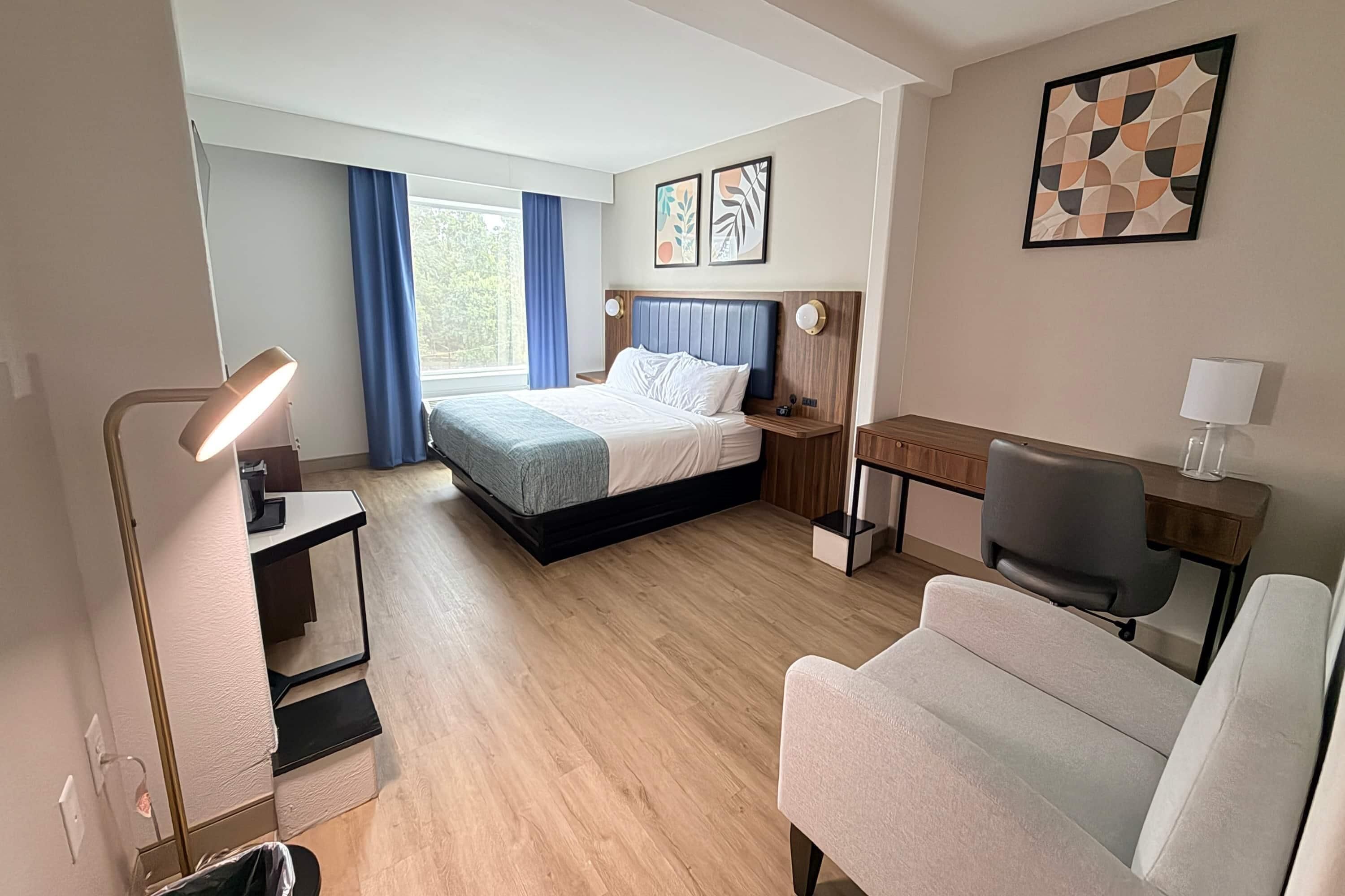 A hotel room with a bed, desk, chair, and a lamp.