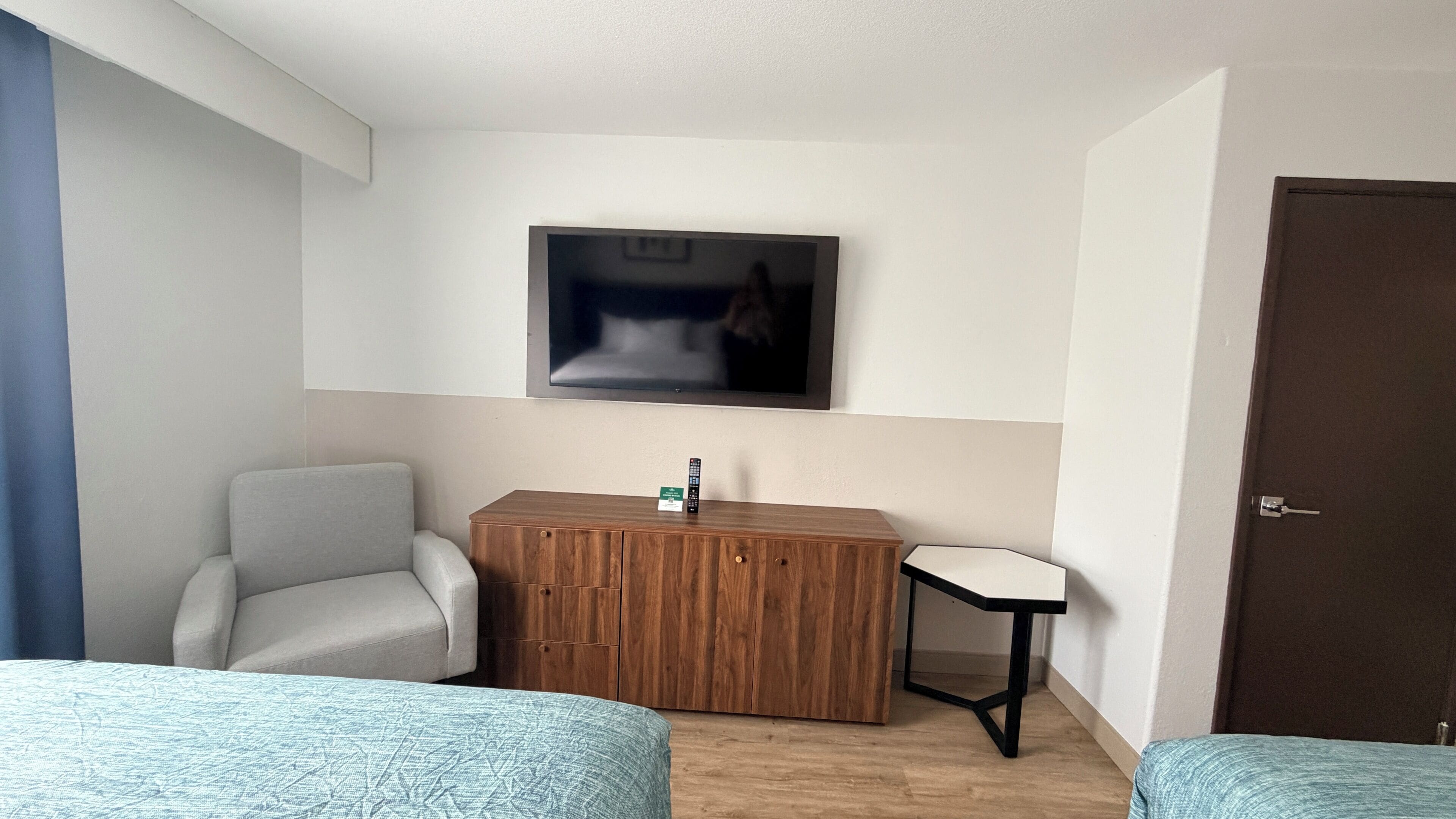 A hotel room with a bed, a desk, a chair, a TV mounted on the wall, and a door.