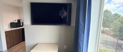 Quarto com televisão de tela plana na parede, um pequeno banco, micro-ondas e janela com vista para o exterior.