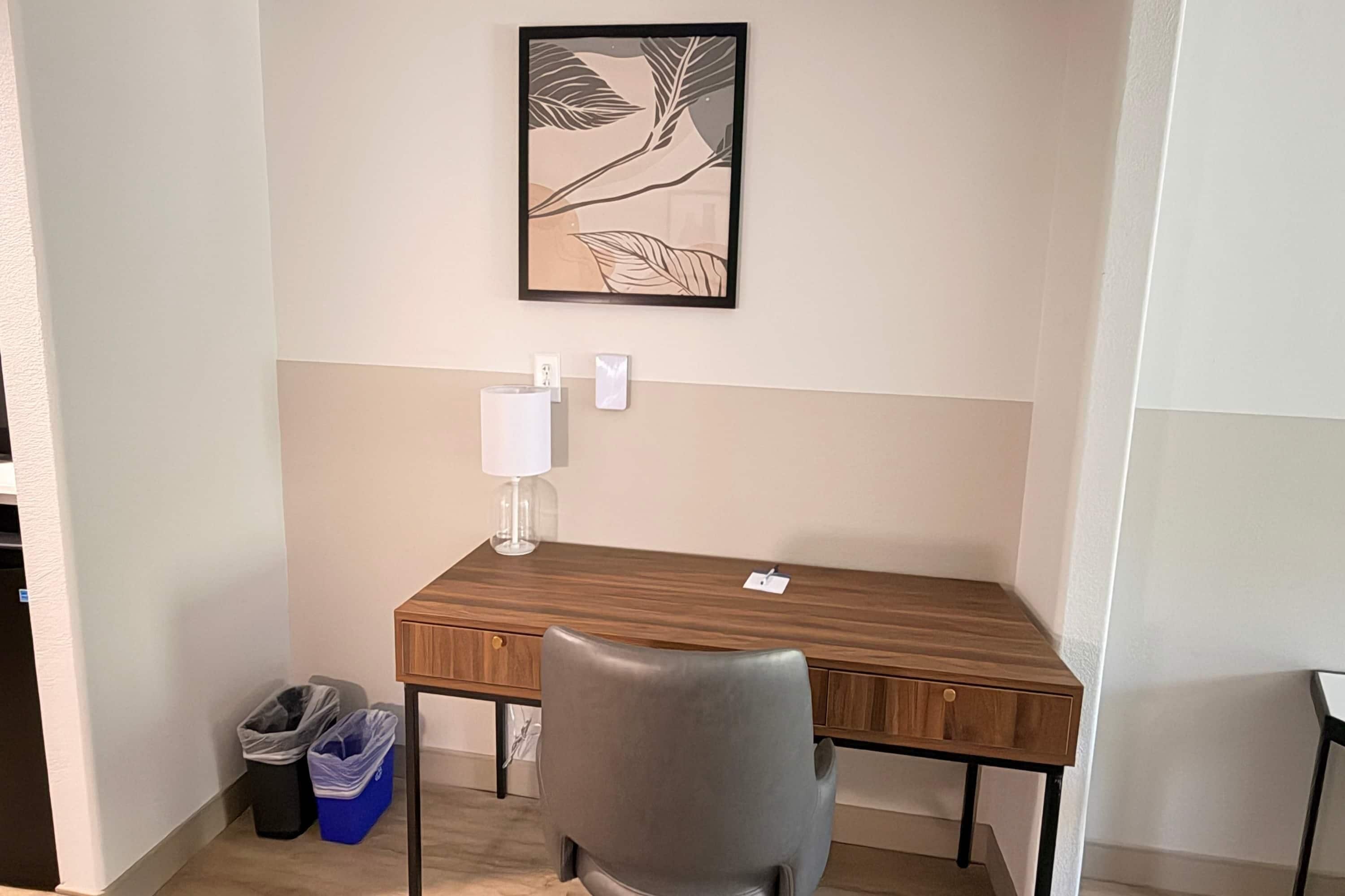 Een compact, verzorgd werkplek met een houten bureau, een grijze stoel, een witte lamp en een kunstwerk in een lijst aan de muur.