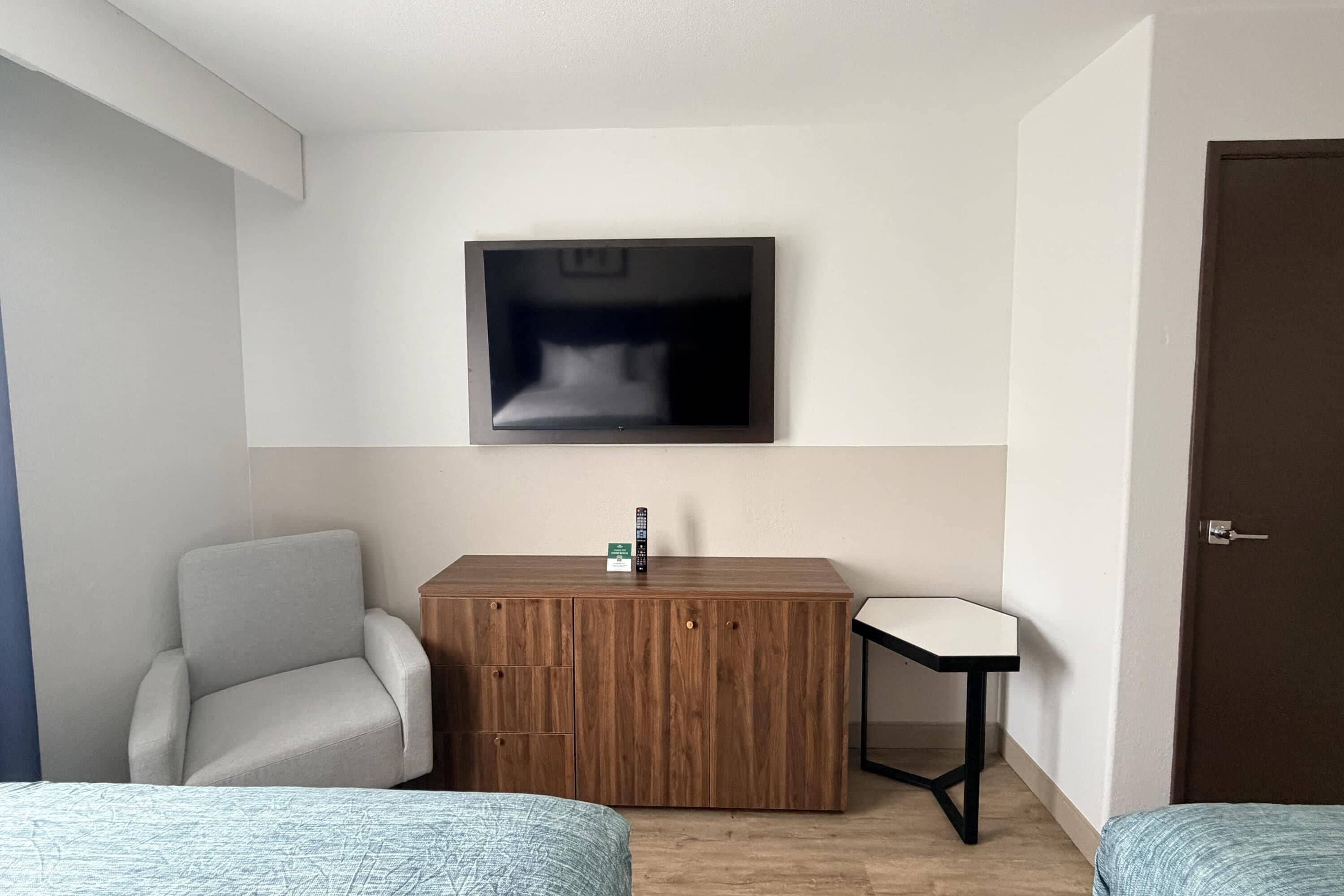 A hotel room with a bed, a desk, a chair, a TV mounted on the wall, and a door.