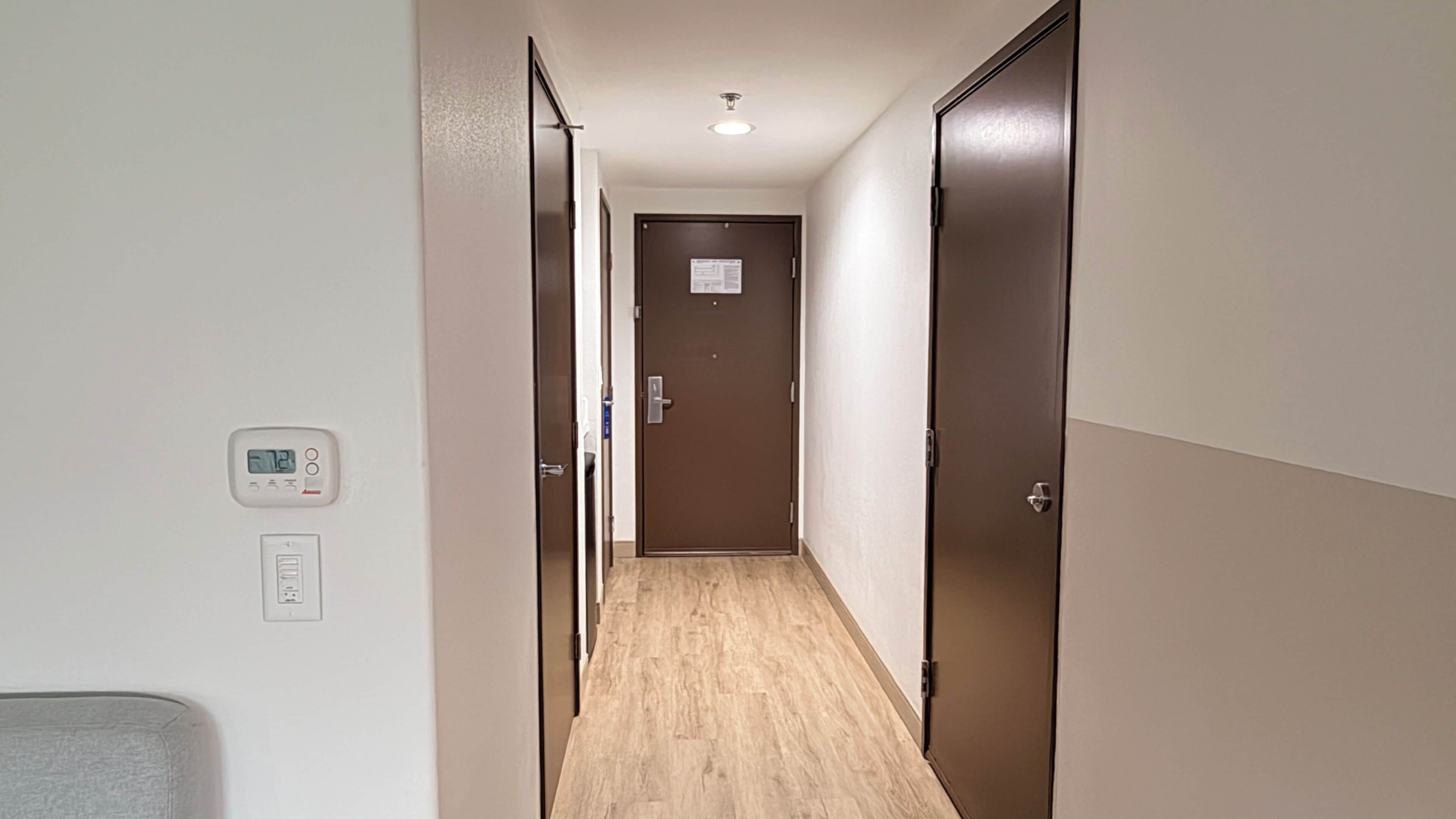 A narrow hallway with wooden flooring, a thermostat on the wall, and a door at the end.