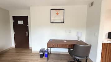 A room with a wooden desk, a chair, a door, a trash bin, and a framed artwork on the wall.