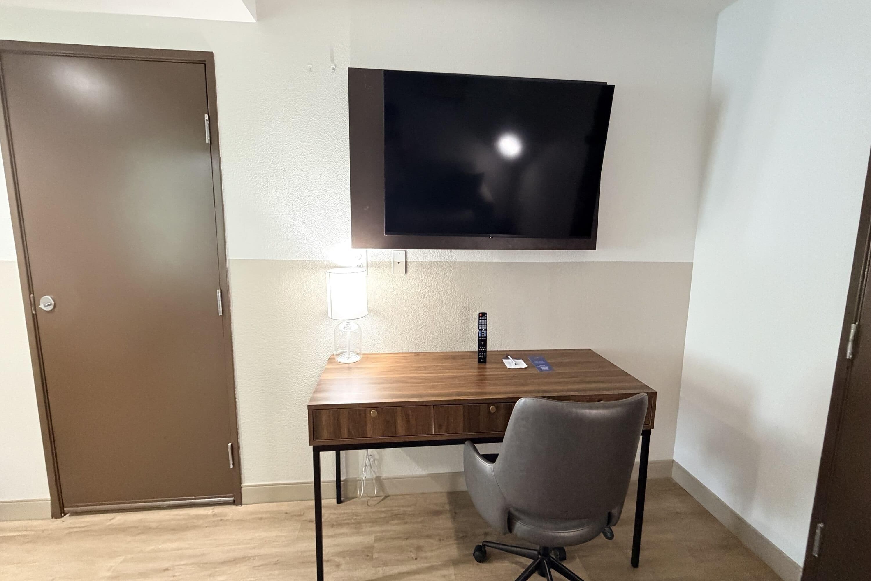 A room with a wooden desk, a grey office chair, a mounted flat-screen TV, and a closed door.