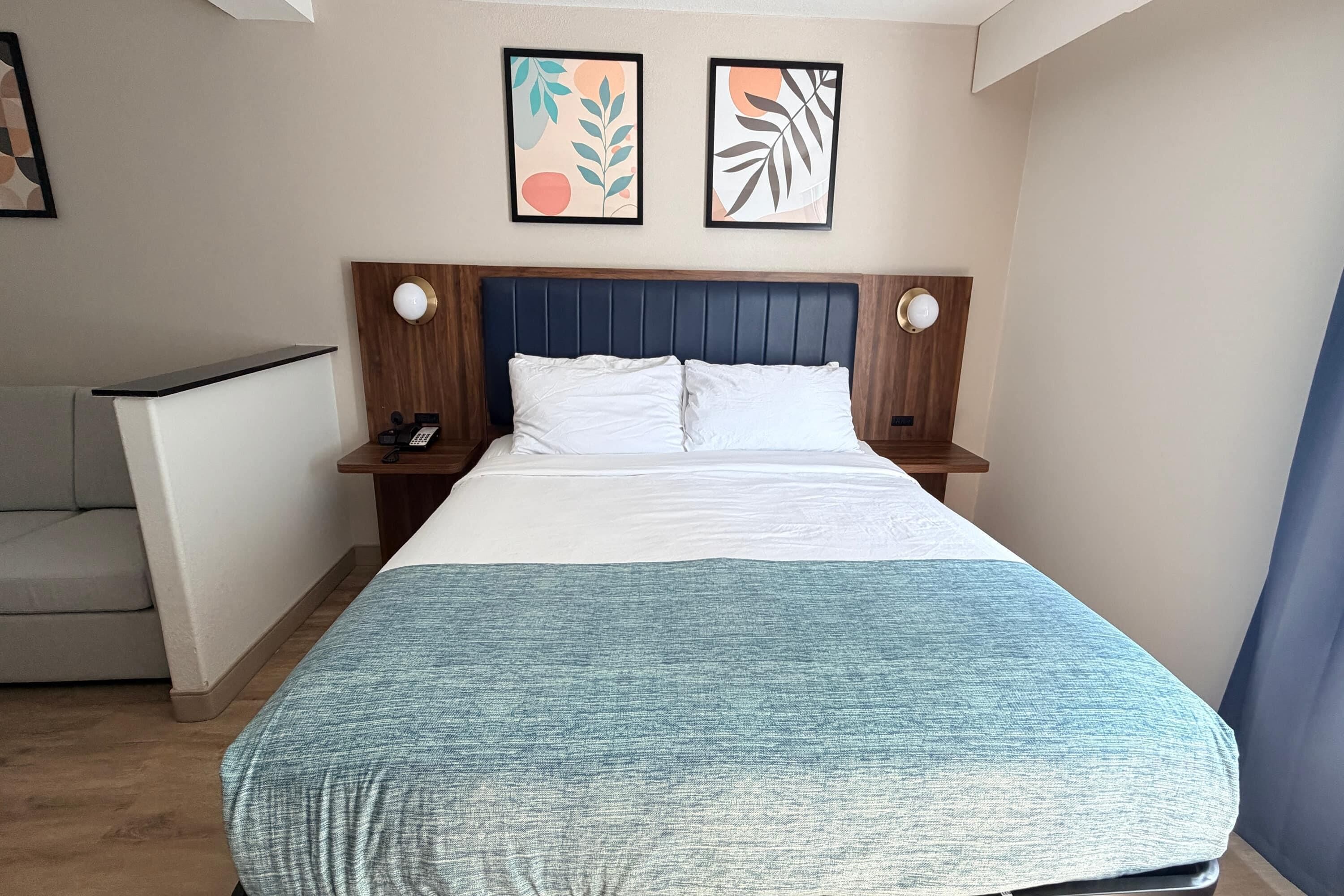 A hotel room with a bed, bedside tables, a desk, and artwork on the wall.