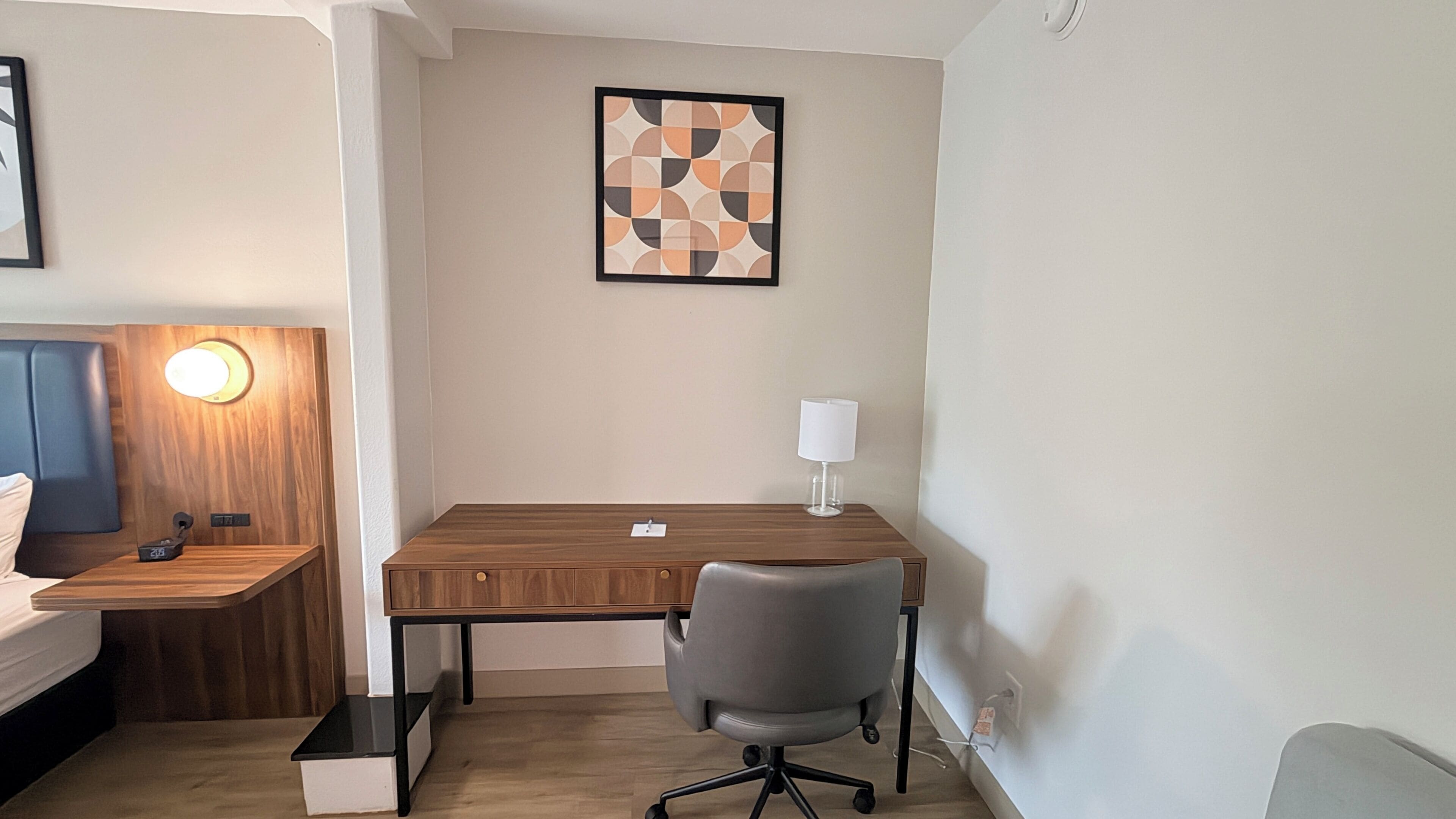 A hotel room with a wooden desk, a grey office chair, a bedside table with a lamp, and a framed abstract artwork on the wall.