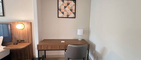 A hotel room with a wooden desk, a grey office chair, a bedside table with a lamp, and a framed abstract artwork on the wall.