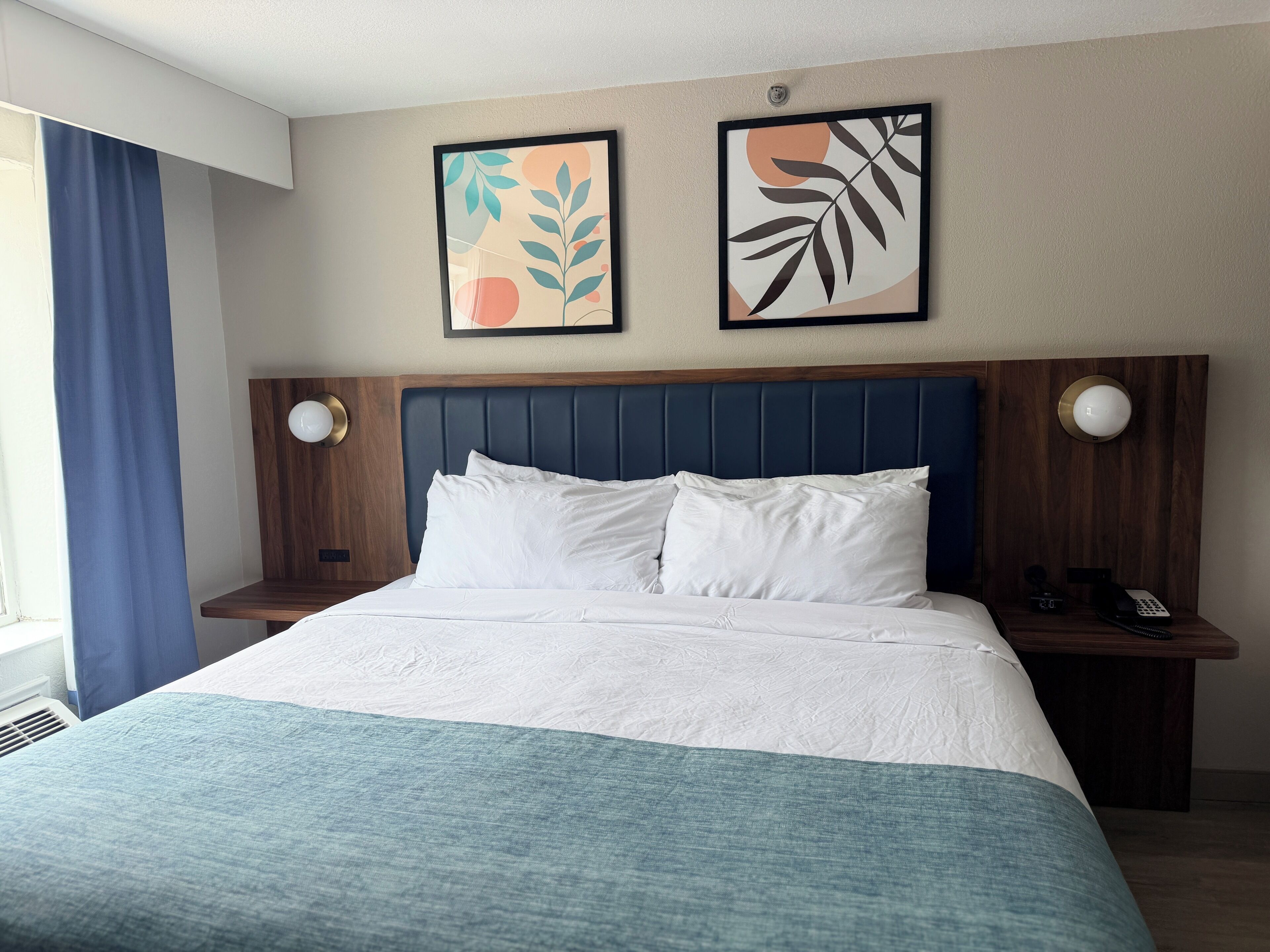 A hotel room with a bed, two wall-mounted artworks, a wooden headboard, bedside tables with lamps, and a window with curtains.