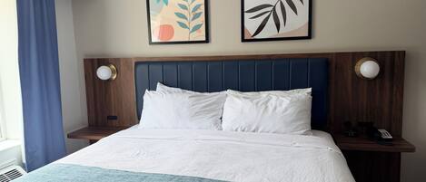 A hotel room with a bed, two wall-mounted artworks, a wooden headboard, bedside tables with lamps, and a window with curtains.