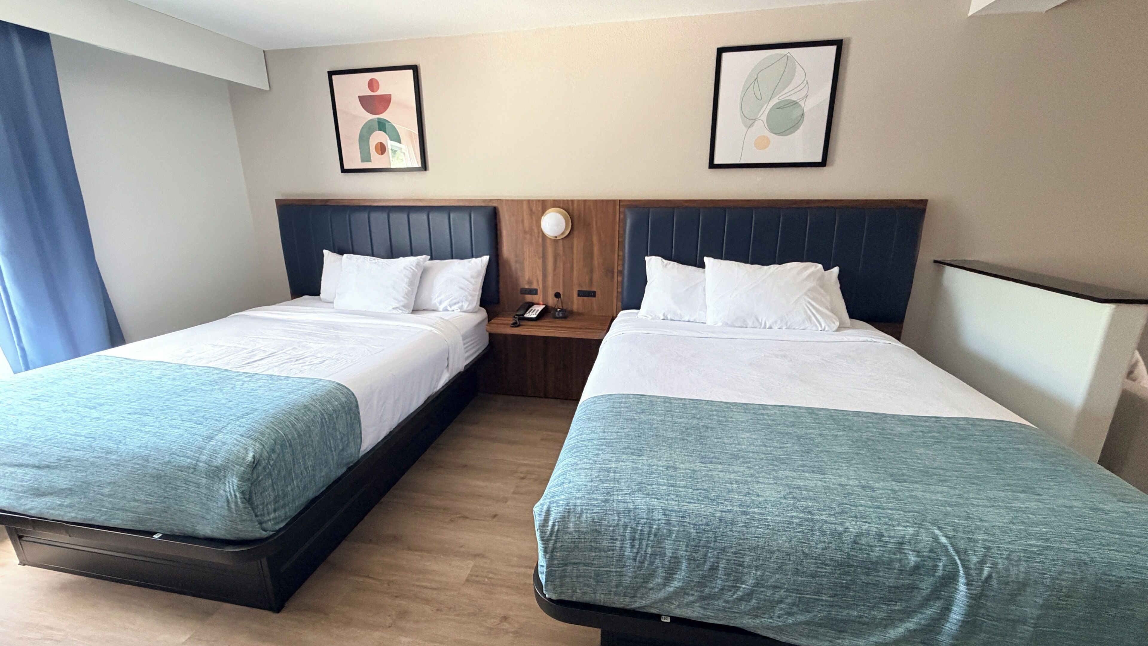 Une chambre d’hôtel avec deux lits, une tête de lit en bois, une table de chevet avec un téléphone et deux œuvres d’art encadrées accrochées au mur.