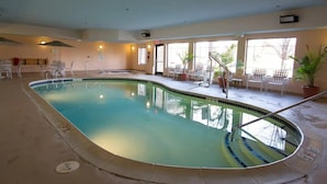 An indoor swimming pool with clear water, surrounded by chairs and potted plants, with large windows allowing natural light.