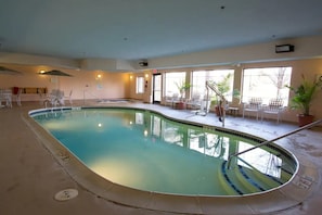 Una piscina interior con agua cristalina, rodeada de sillas y plantas en macetas, con ventanas amplias que dejan entrar luz natural.