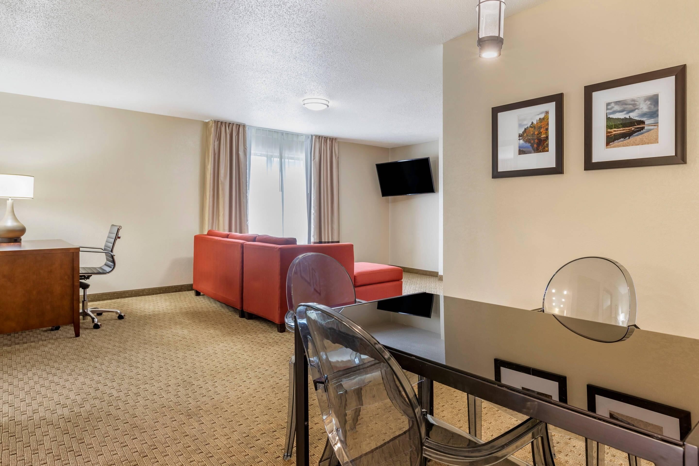 A hotel room with a dining table, a red sofa, a clear chair, a lamp, a TV, and two framed pictures on the wall.