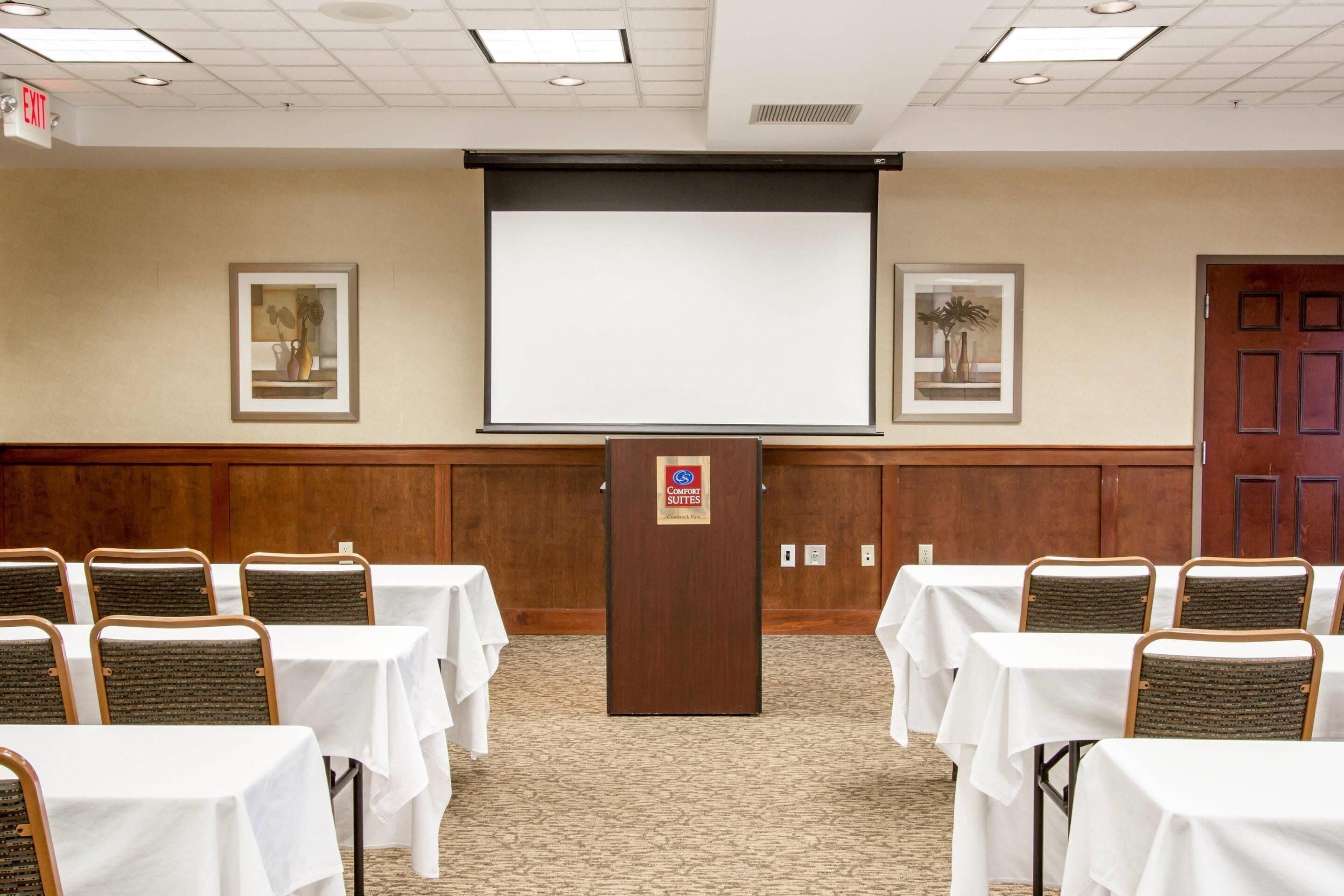 A conference room with a projector screen, tables, chairs, and framed artwork on the walls.