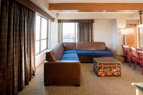A hotel room with a sectional sofa, a small ottoman, a wooden chest, and a dining area with red chairs. - Great Wolf Lodge Bloomington (Bloomington)