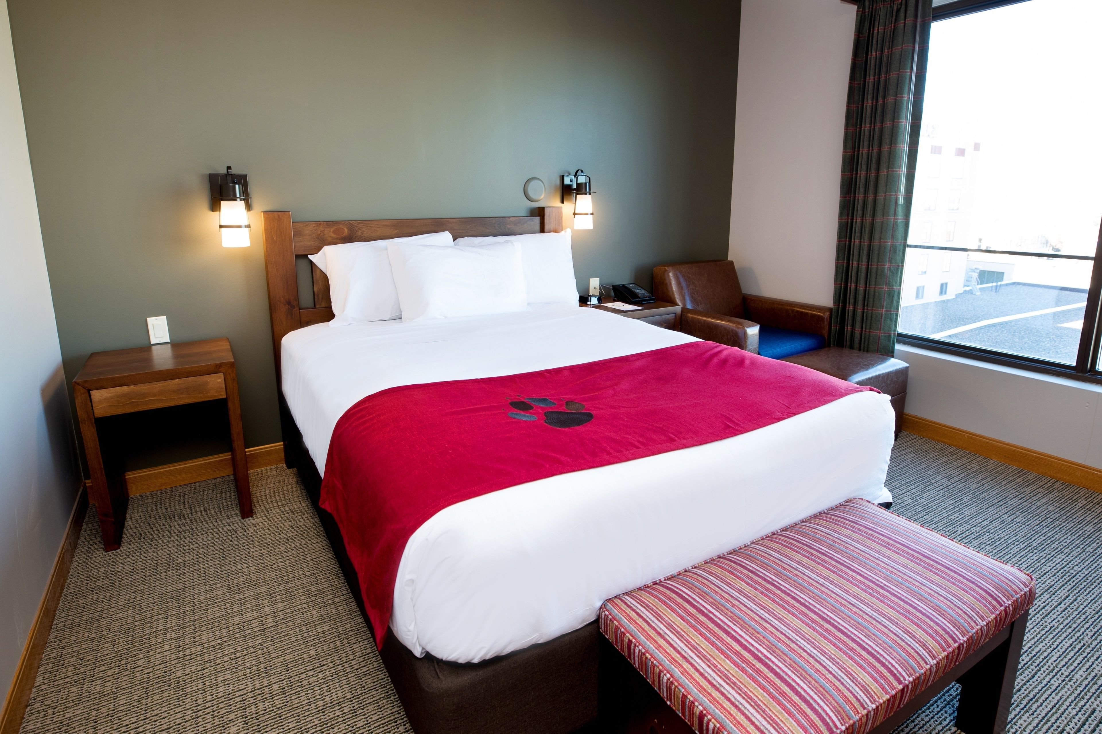 Habitación de hotel con una cama grande, cabecera de madera, dos mesitas de noche, una manta roja, un banco a rayas y vista a la calle por la ventana.
