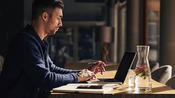 Una persona trabajando en una mesa de madera con una computadora portátil, un cuaderno y un vaso con una planta.