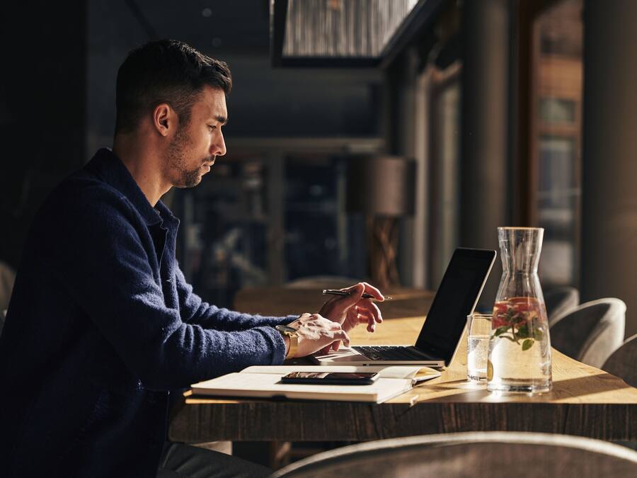 Una persona trabajando en una mesa de madera con una computadora portátil, un cuaderno y un vaso con una planta.