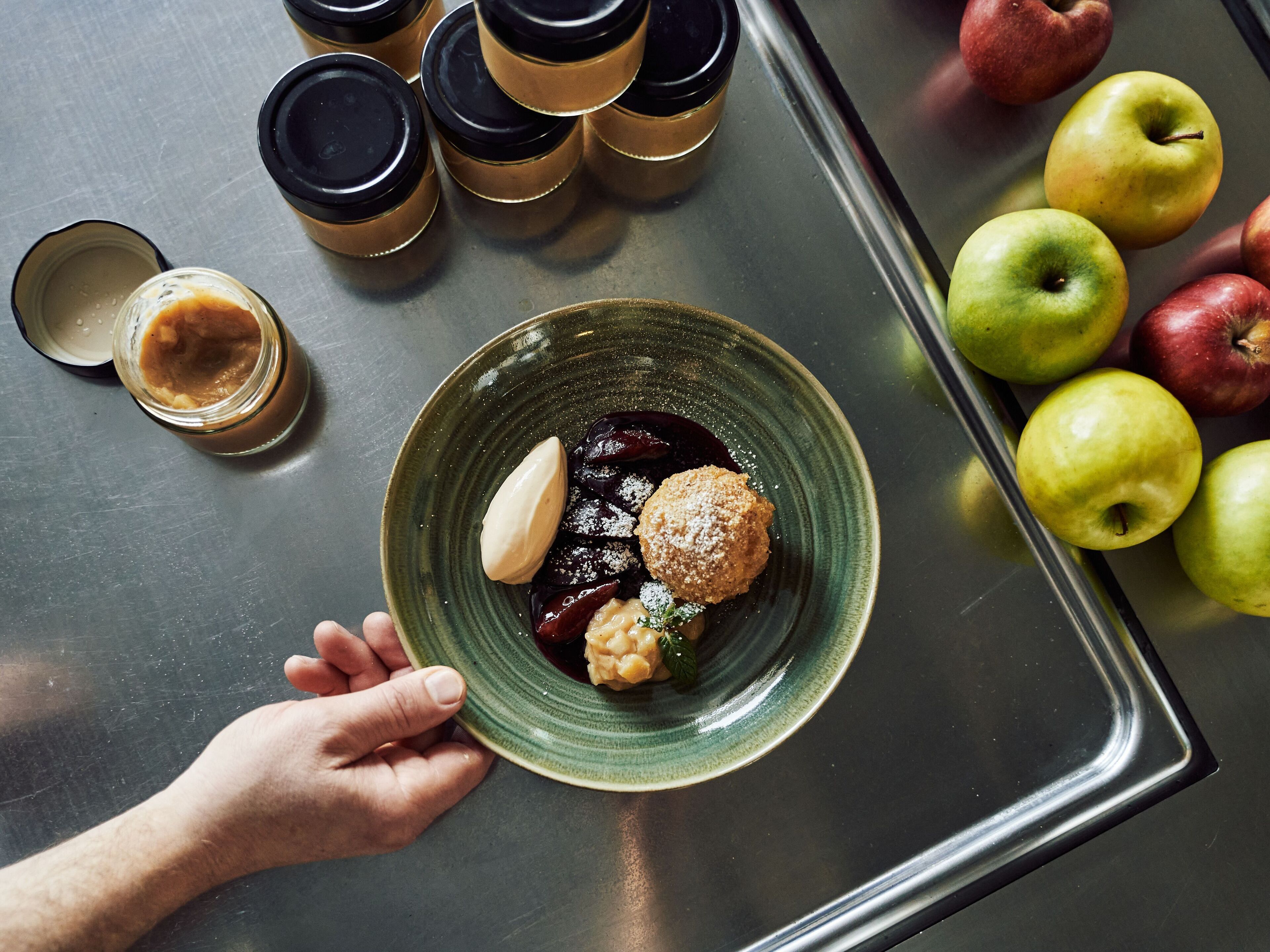 A hand holding a plate with a dessert, surrounded by apples and coffee cups on a metallic tray.