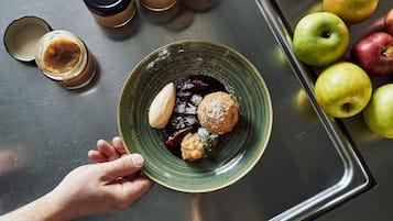 A hand holding a plate with a dessert, surrounded by apples and coffee cups on a metallic tray.