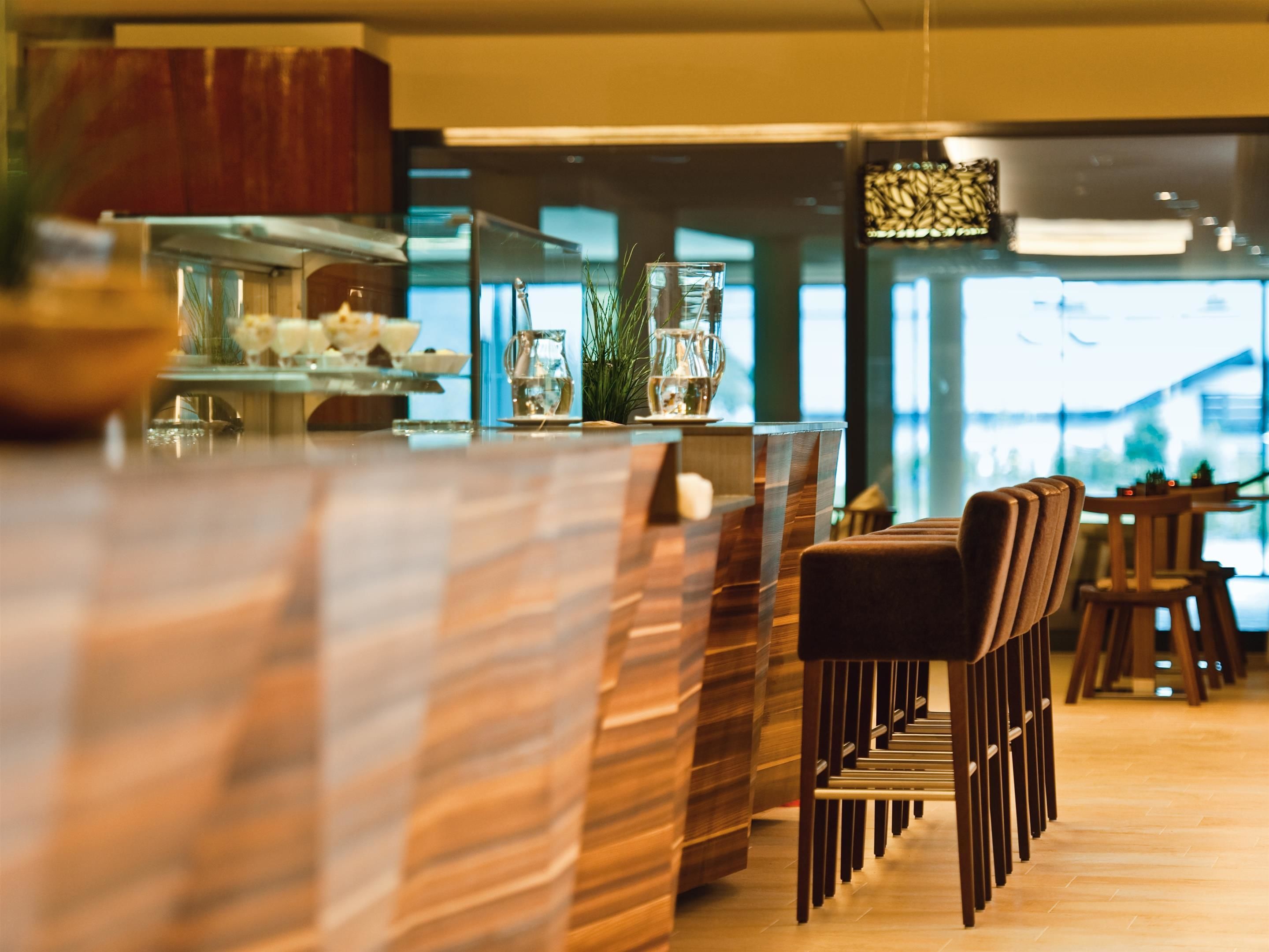 A modern bar area with a wooden bar counter, seating with cushioned chairs, and a large window providing natural light.