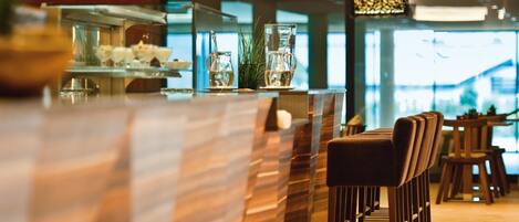 A modern bar area with a wooden bar counter, seating with cushioned chairs, and a large window providing natural light.