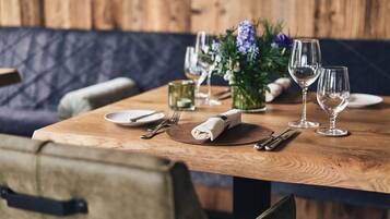 A wooden dining table set with plates, cutlery, and wine glasses, with a floral centerpiece and a cushioned bench in the background.