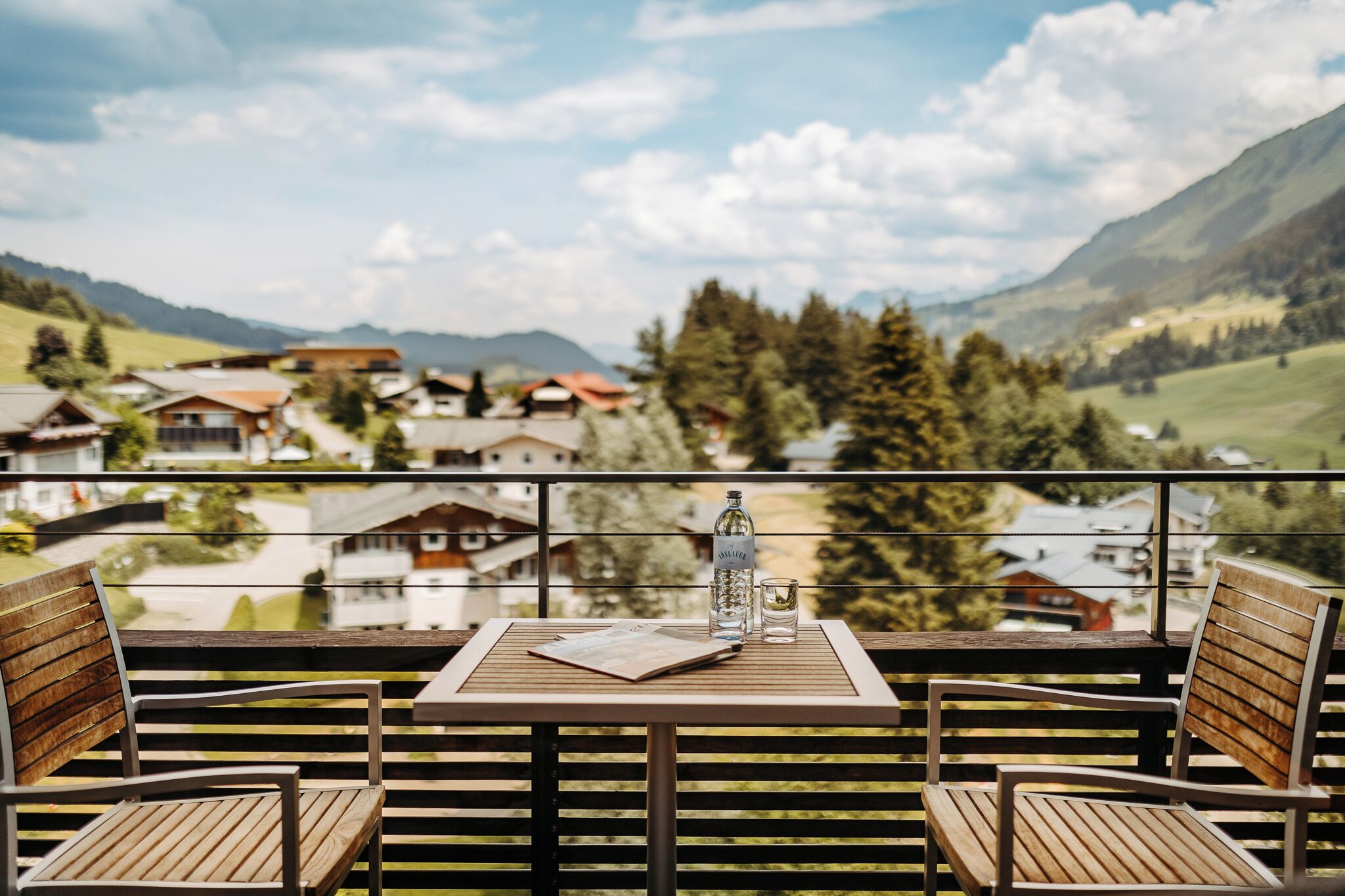 Un balcón con sillas y una mesa de madera, con vistas a un pueblo con casas y montañas.
