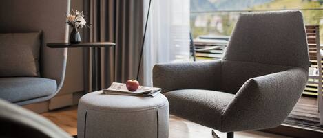 Un salon moderne avec un fauteuil gris, un petit pouf sur lequel reposent un livre et une pomme, une table d’appoint ornée d’un vase de fleurs, et une vue sur les montagnes depuis un balcon.