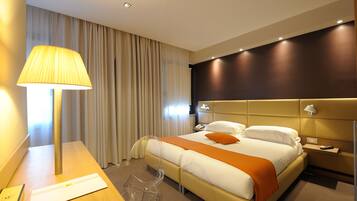 A hotel room with a bed, a desk with a lamp, a chair, and a bedside table.