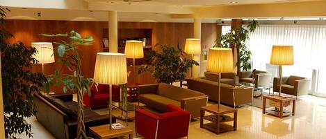 Un moderno vestíbulo de hotel con un área de estar central, varias lámparas y plantas en macetas.