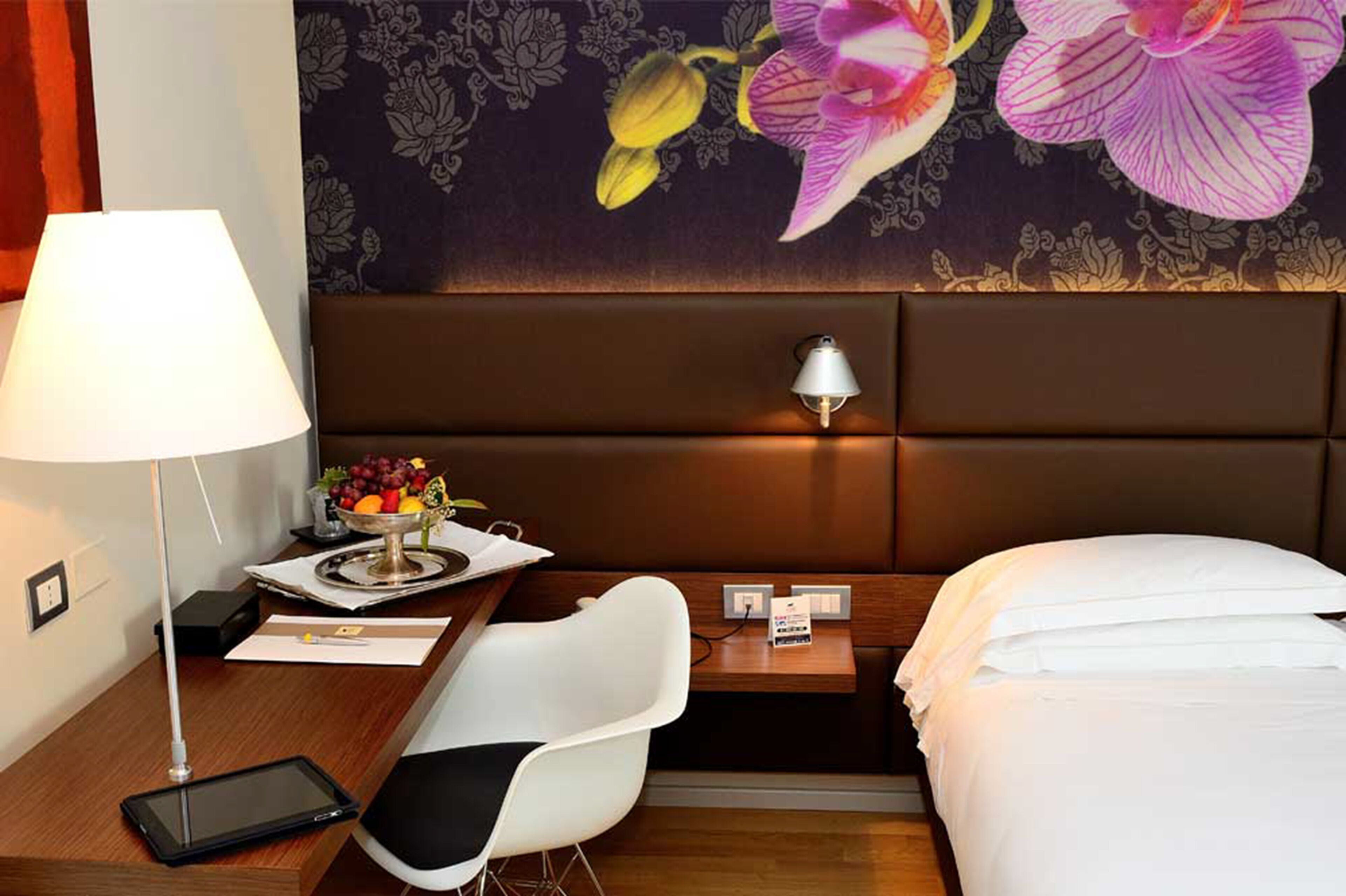 A hotel room with a bed, a desk with a lamp, a chair, a bedside table with a lamp, and a wall with a floral wallpaper.