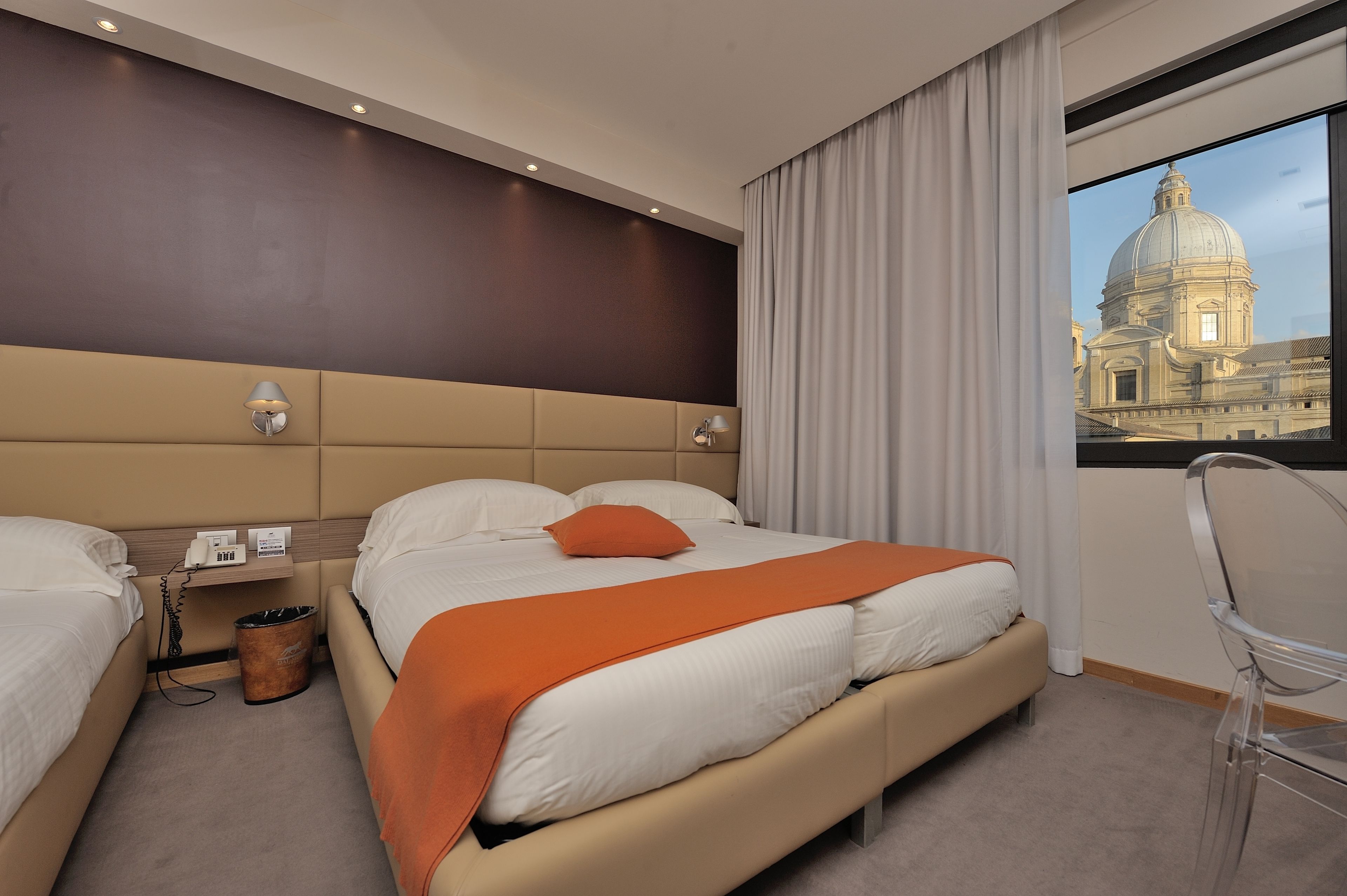Une chambre d’hôtel avec un grand lit, une table de chevet avec un téléphone et une poubelle, une chaise et une fenêtre donnant sur un immeuble.