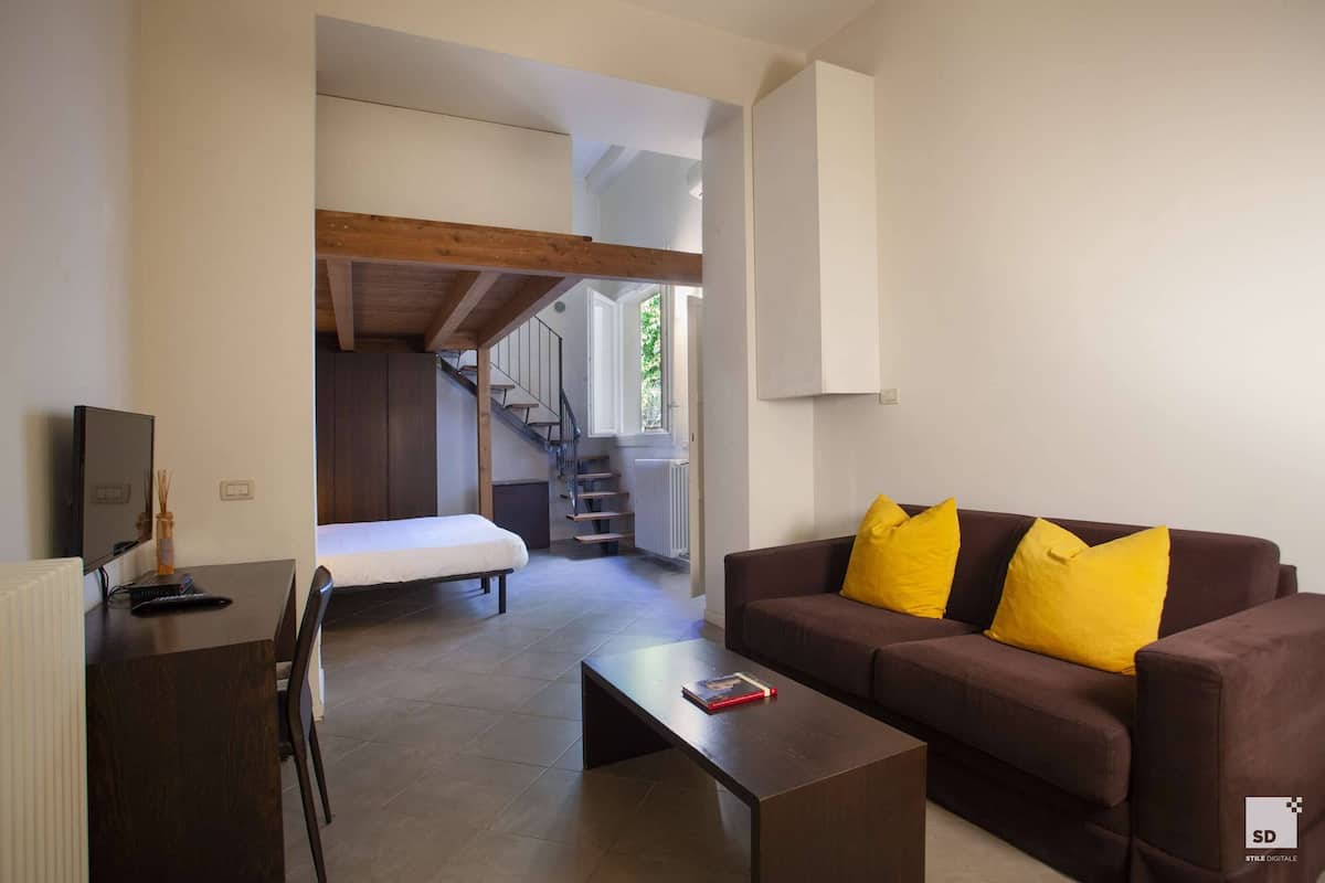 Ein modernes Wohnzimmer mit einer braunen Couch, einem Couchtisch und einem Fernseher auf einem Holztisch. Eine Treppe führt auf eine höhere Ebene.