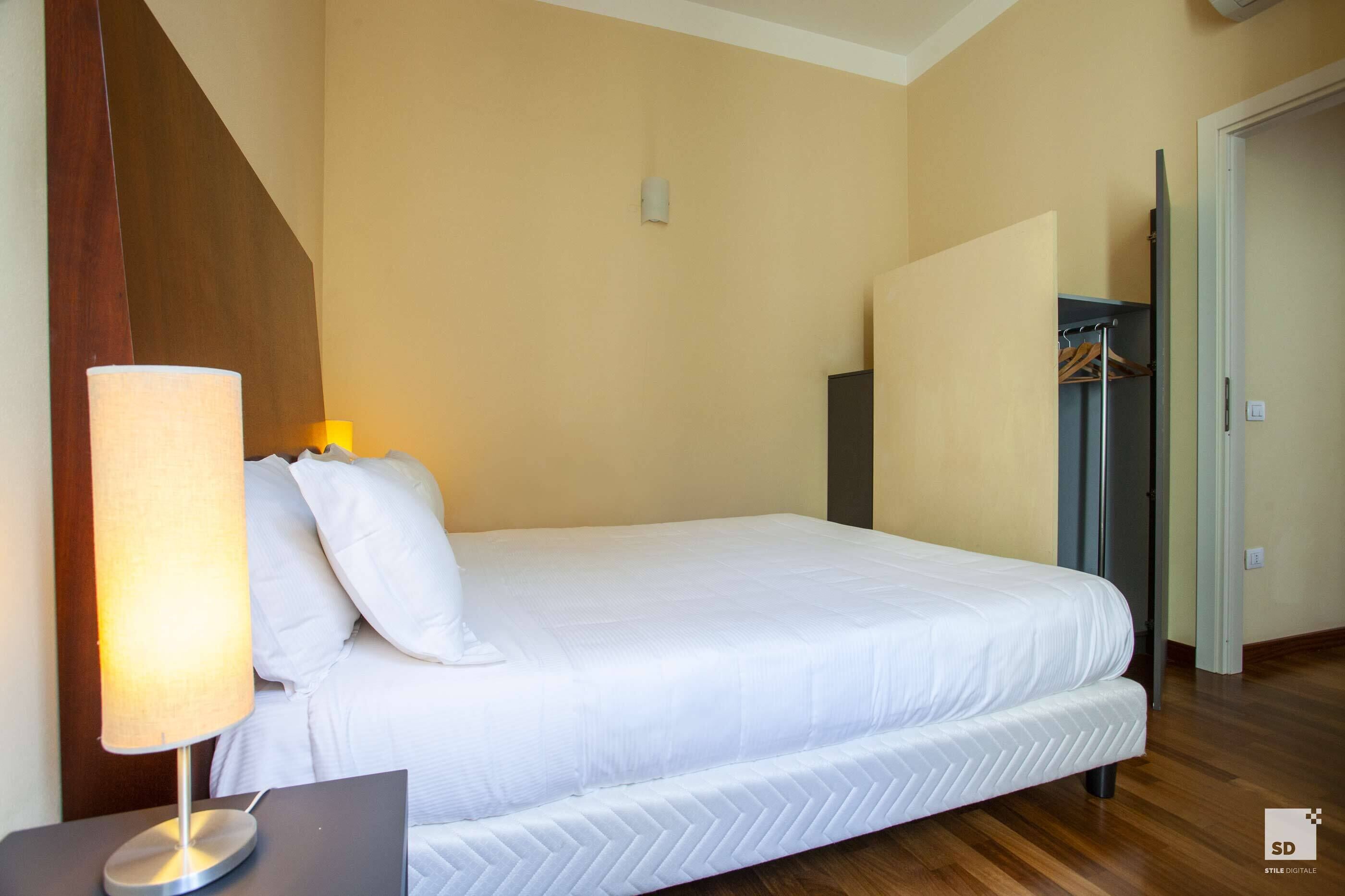 A neatly made bed with a white comforter and pillows, a bedside lamp, and a wooden floor.